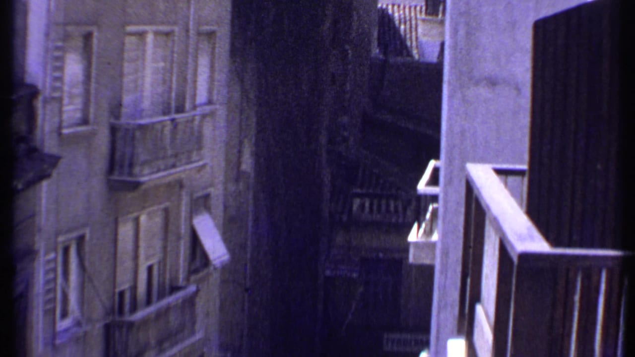 1969 Kenya home-movie frame looking down between concrete residential buildings with balconies, shutters, awnings, and deep shadow.