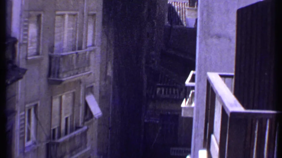 1969 Kenya home-movie frame looking down between concrete residential buildings with balconies, shutters, awnings, and deep shadow.