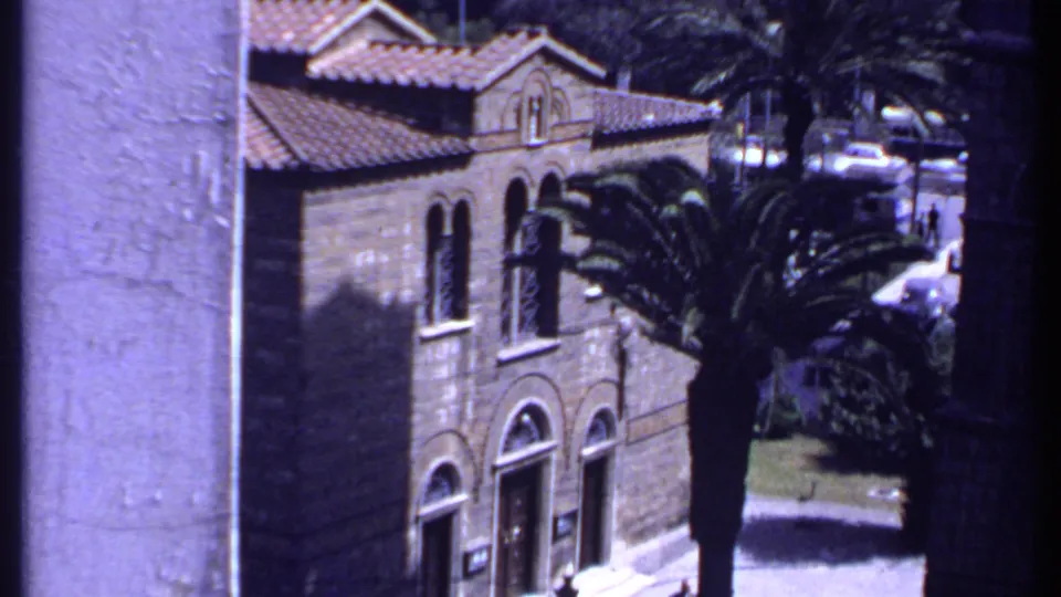 Kenya 1969 home-movie view of an arched masonry building, palm fronds and older cars on a nearby road.