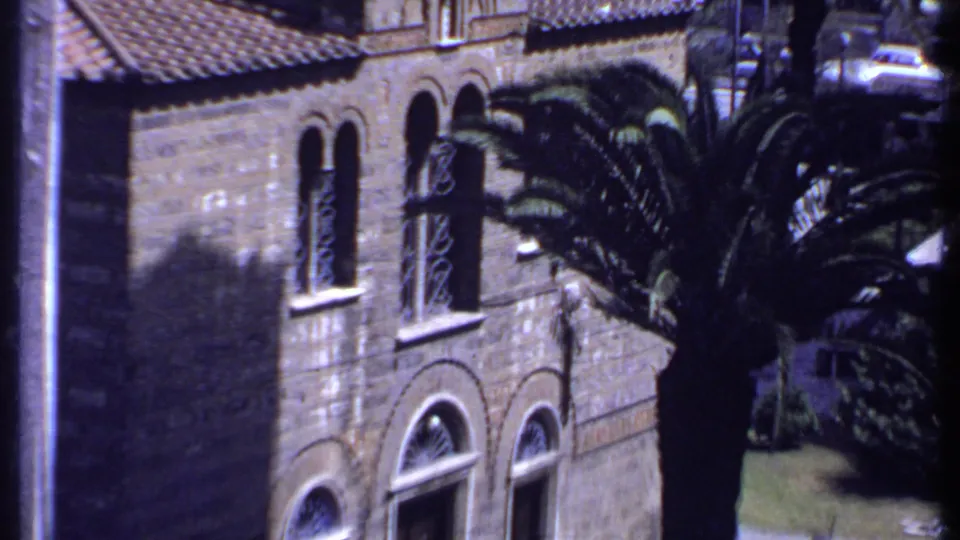 1969 Kenya frame with palm fronds in front of an arched masonry facade and cars along the road.
