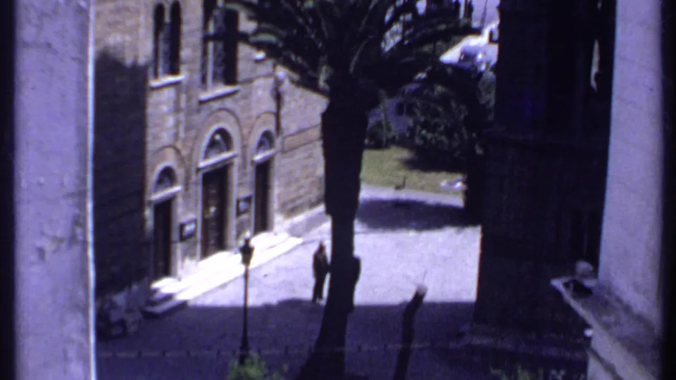 1969 Kenya home-movie frame looking down at the church courtyard, with a palm, arched facade, and a small figure walking nearby.