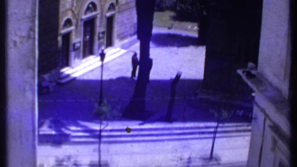 1969 Kenya home-movie frame showing a stone building facade, steps, shadows, and a small figure walking below.