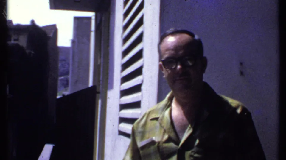 Adult man in a green shirt and glasses stands near a white louvered shutter in 1969 Kenya home-movie footage.