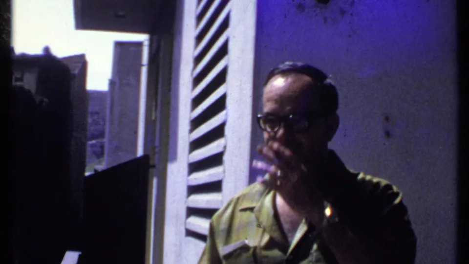 Adult man in a green patterned shirt raises his hand toward his mouth near a shuttered doorway in 1969 Kenya footage.