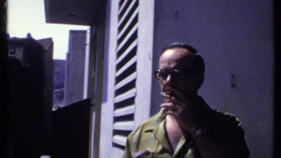 Adult man in glasses remains near the white louvered shutter, hand at his mouth, in a softly exposed 1969 Kenya frame.