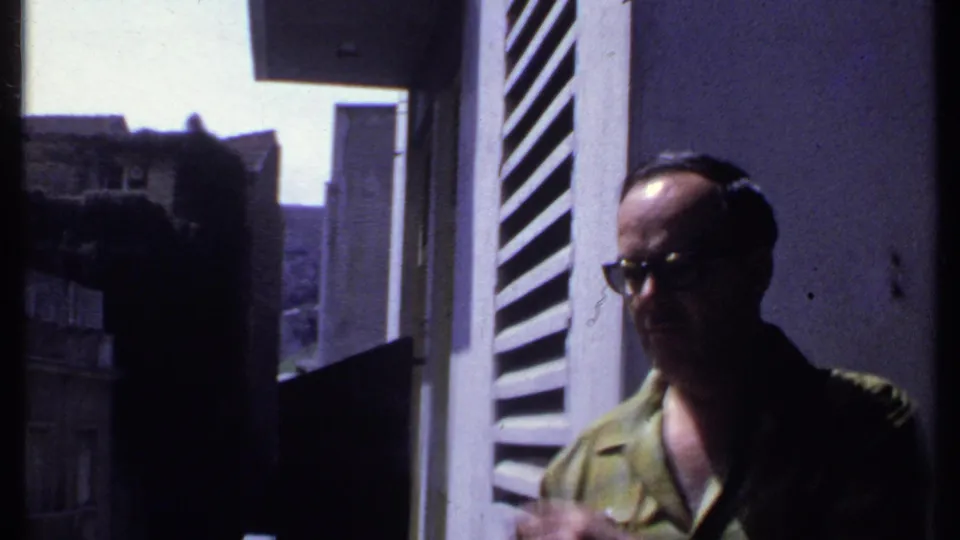 In Kenya 1969 footage, an adult man in glasses stands on a balcony with open shutters and city buildings behind him.