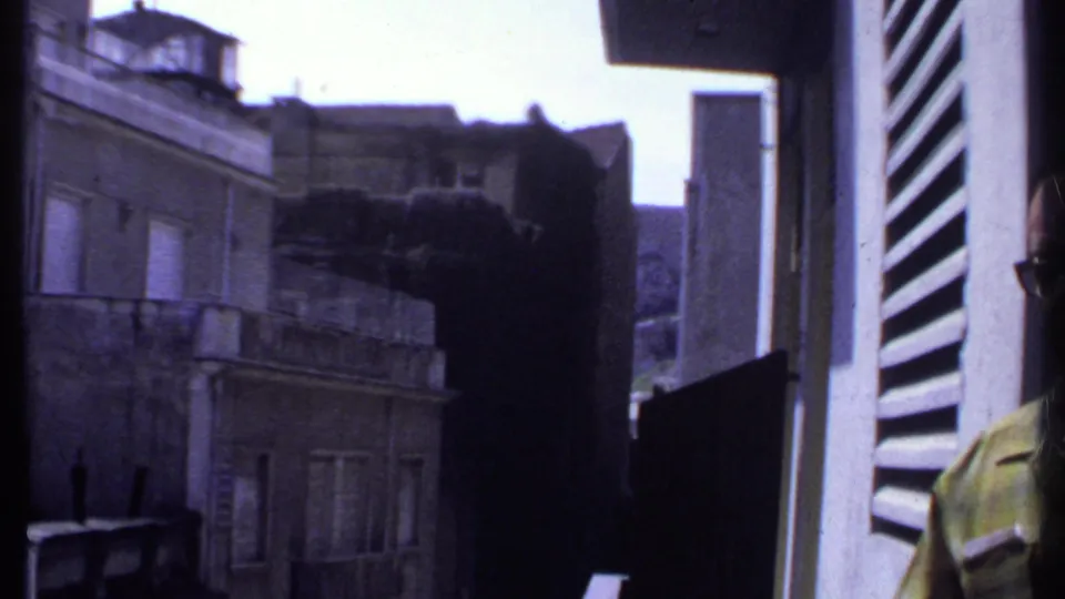 Kenya 1969 home-movie frame looking past balcony shutters toward dense masonry buildings and a rooftop structure.