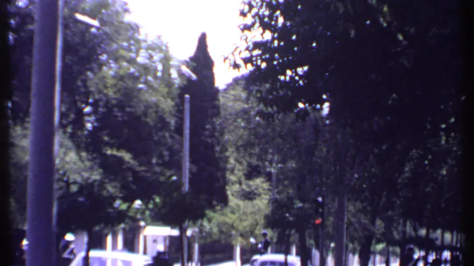 1969 Kenya home-movie frame showing a shaded street with stone walls, tall trees, a roadside pole, and a classic car.