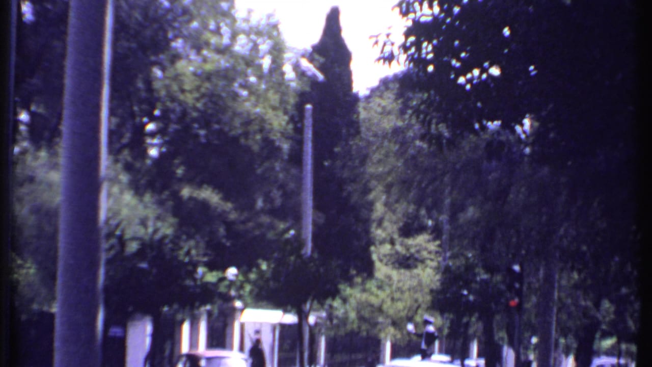 Shaded roadside view with trees, stone walls, a small building, cars, and distant pedestrians.