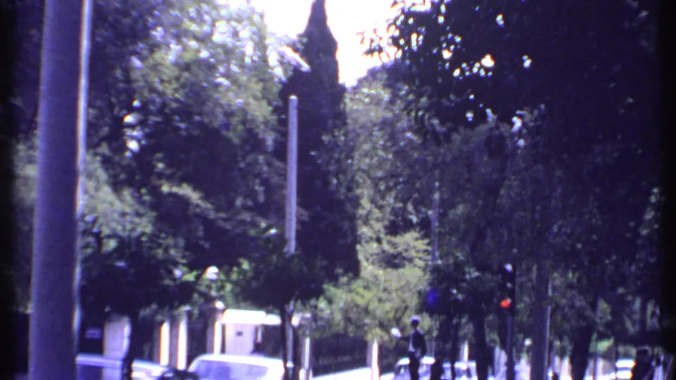 1969 Kenya home-movie frame with a tree-lined street, stone wall, classic cars, pedestrians, and a red traffic signal.