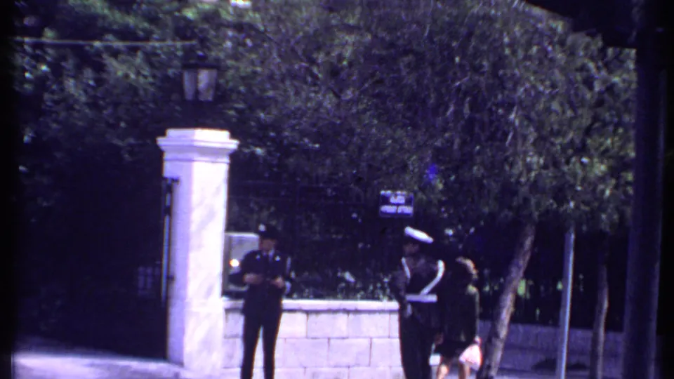 1969 Kenya street frame showing uniformed officers near a white gate pillar, stone wall, and shaded trees.