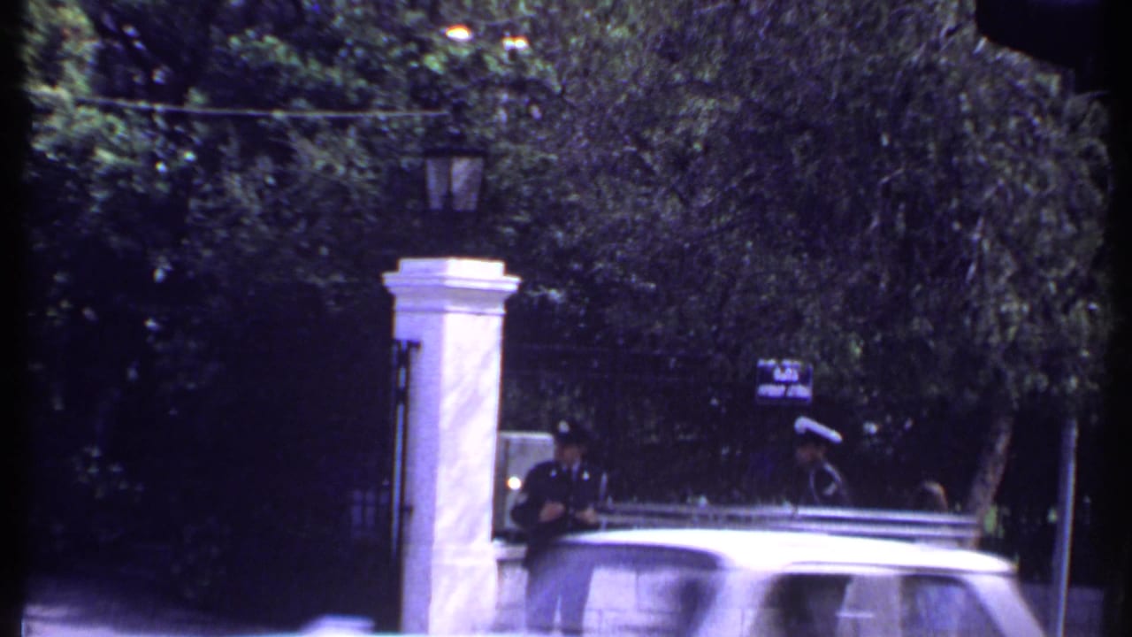 Uniformed guard near white gatepost as a car passes in front of a leafy entrance.