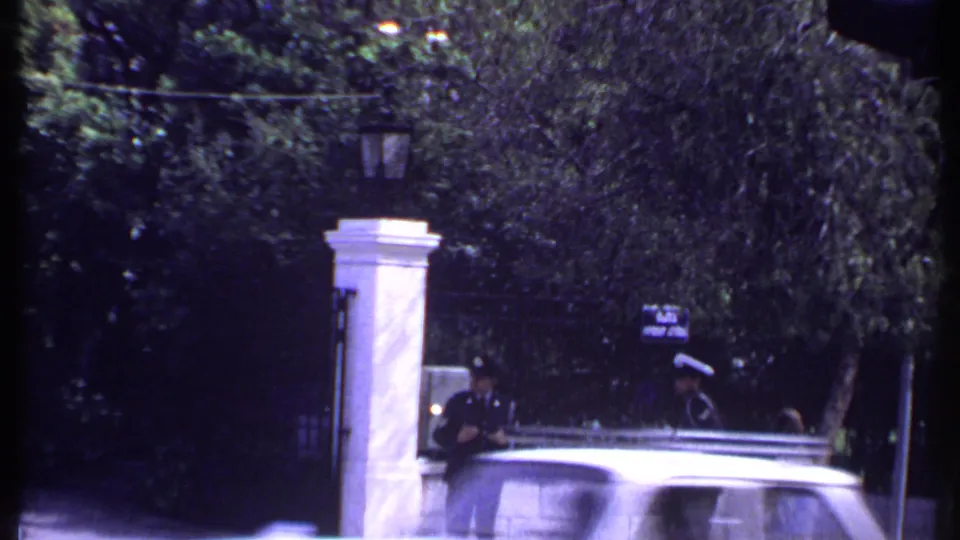 1969 Kenya home-movie frame showing a uniformed guard at a white stone gate while a car passes across the foreground.