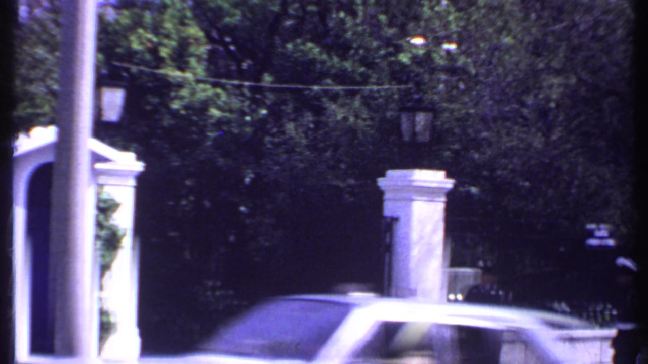 Blurred car passes a gated entrance with a guard partly visible near the right edge.