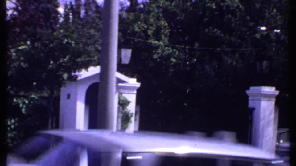 1969 Kenya roadside view of a car moving past a formal gated entrance with stone posts, fencing, lamps, and dense trees.