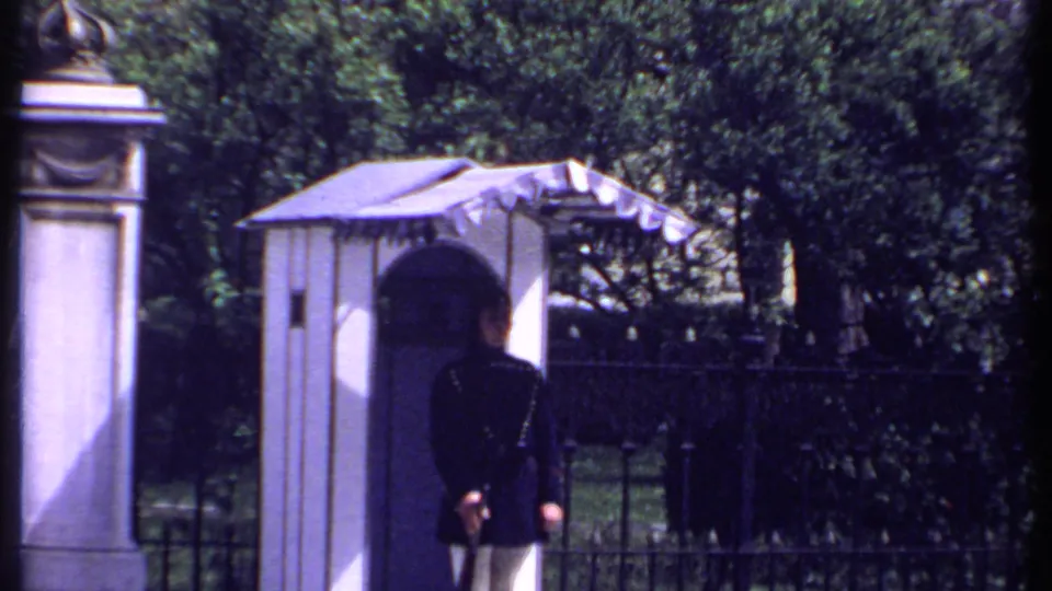 1969 Kenya frame showing a uniformed guard from behind beside an arched shelter, iron fencing, stonework, and trees.