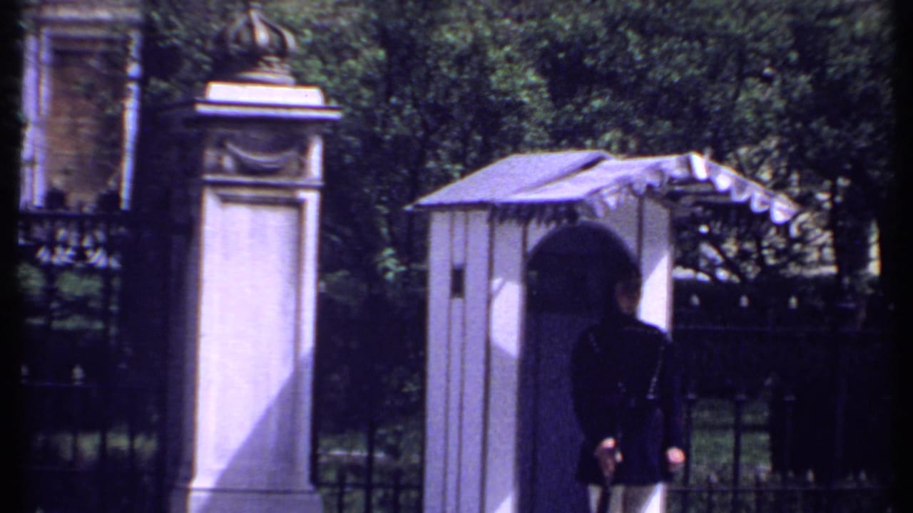 1969 Kenya home-movie frame showing a dark-uniformed guard by a striped guardhouse at ornate gates with crowned stone posts.