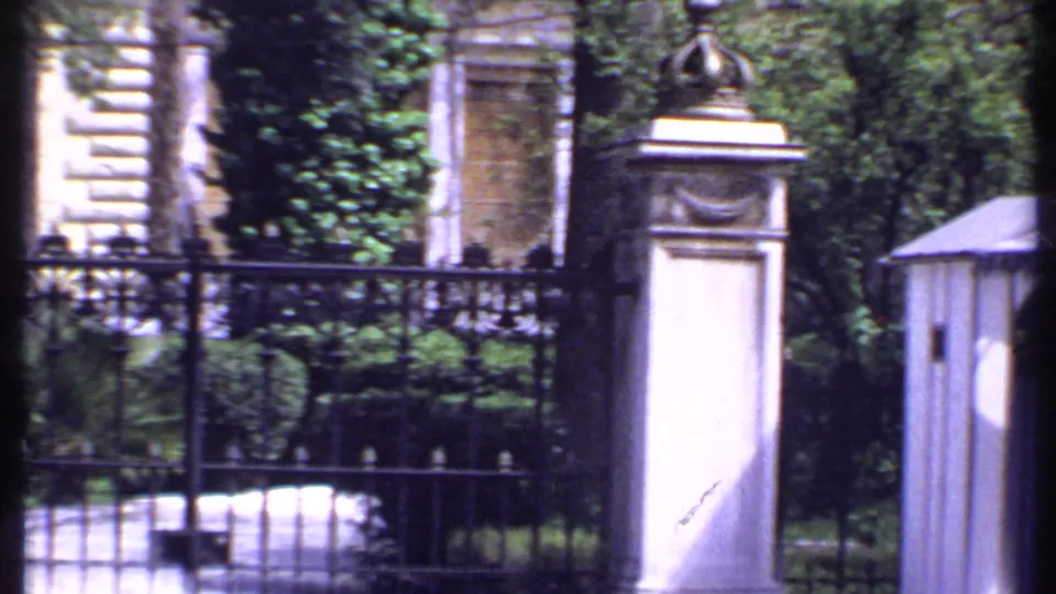 1969 Kenya frame of crowned gateposts and black iron fencing in front of greenery and a pale masonry building.