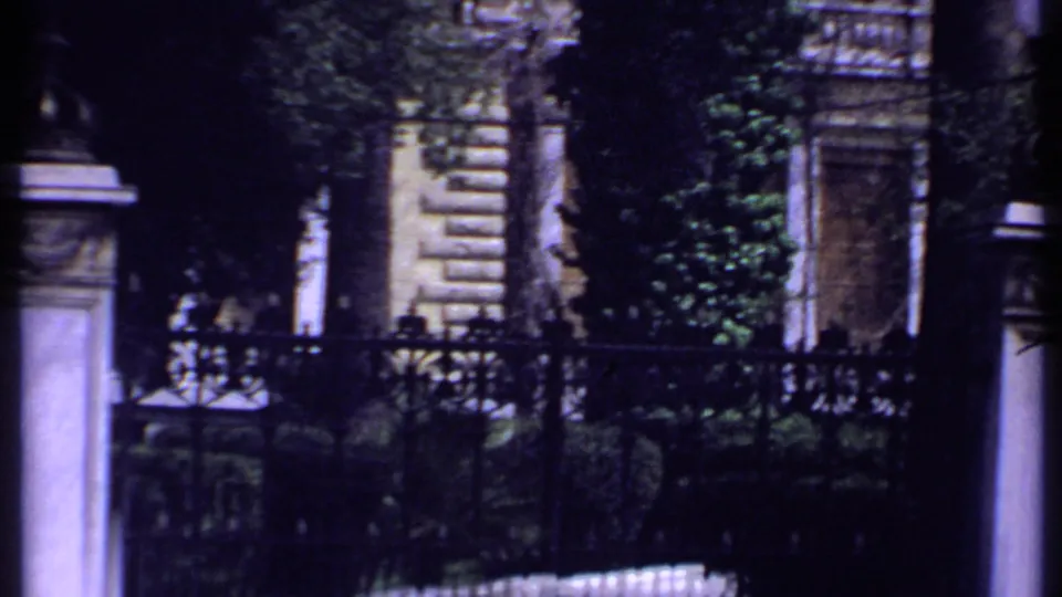 Handheld 1969 Kenya home-movie view of iron fencing, stone gateposts, greenery, and partial building details through a dark foreground band.