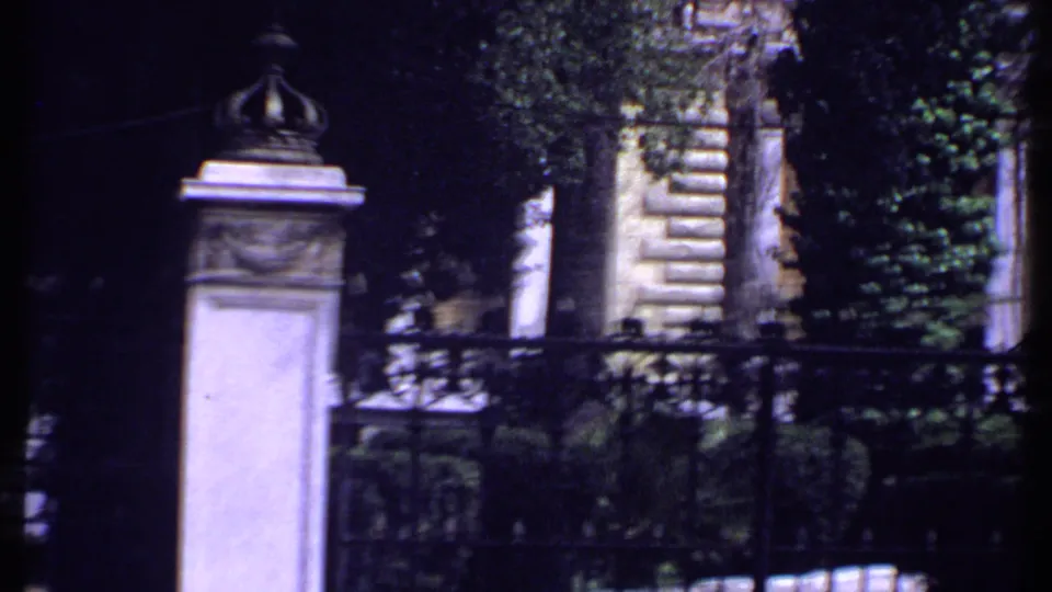 1969 Kenya frame with a crowned stone gatepost, black iron fence, dense greenery, and pale masonry behind the entrance.