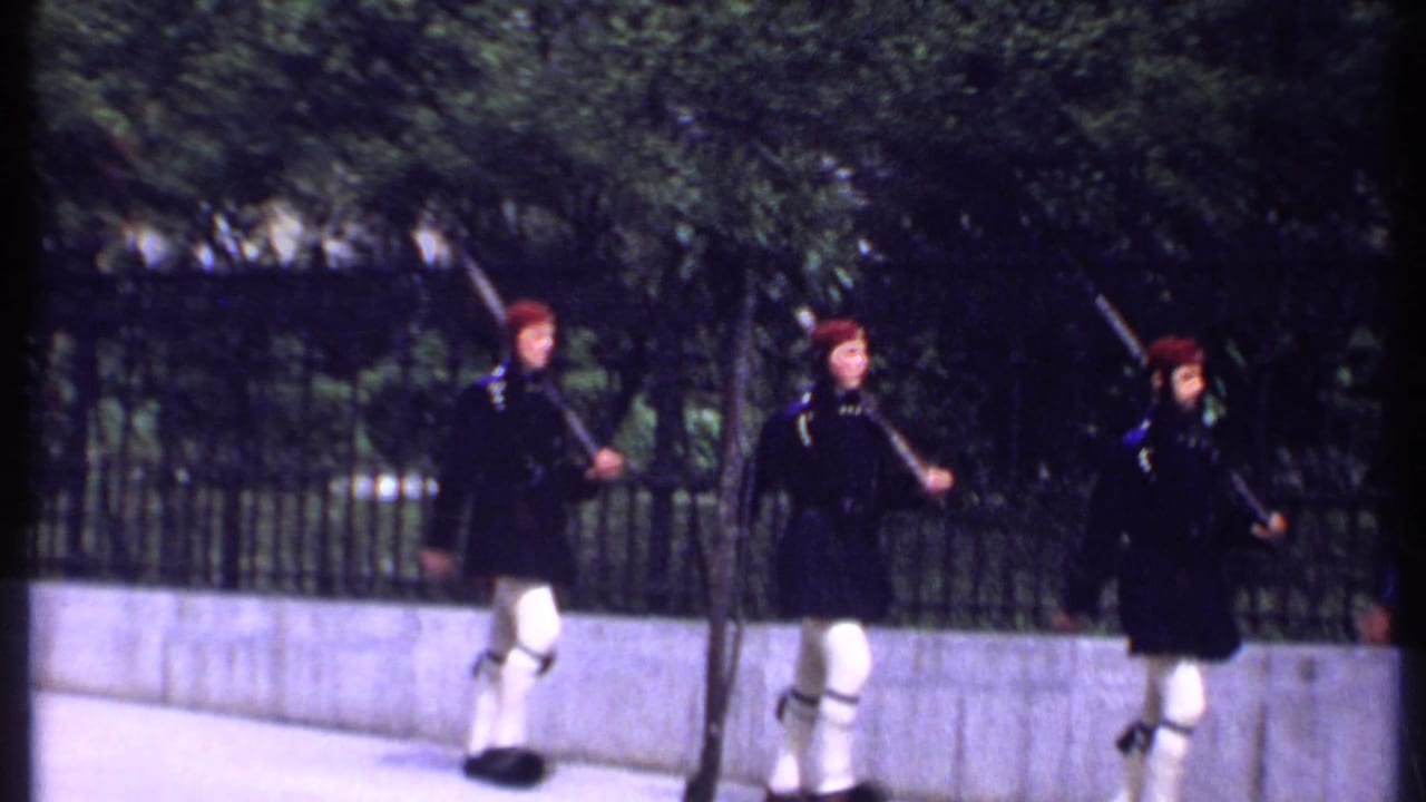 In Kenya 1969 home-movie footage, three uniformed men in red berets march with rifles along a sidewalk by an iron fence.