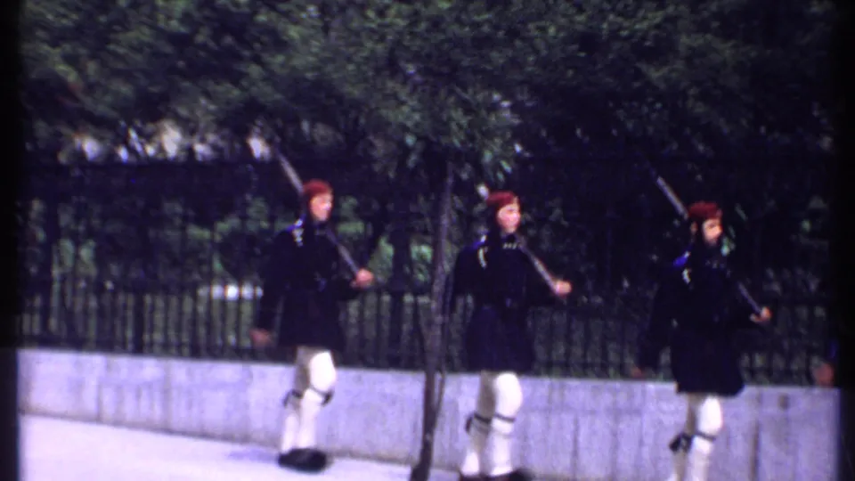 In Kenya 1969 home-movie footage, three uniformed men in red berets march with rifles along a sidewalk by an iron fence.