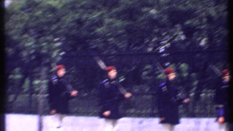 Uniformed men in red berets and dark jackets move across the frame with rifles, with trees and fencing behind them.