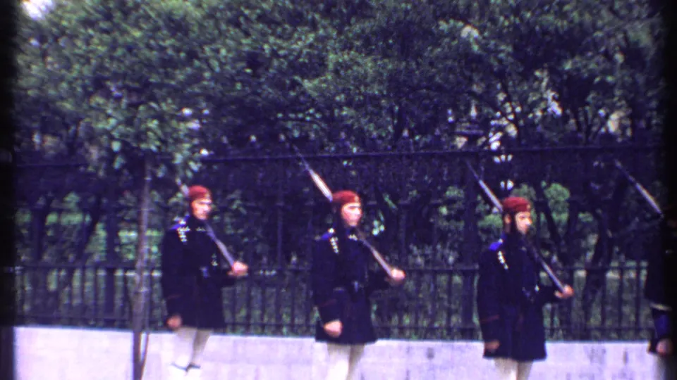 Three uniformed men in red berets, dark jackets, and white trousers march with rifles along a fenced sidewalk in Kenya 1969 footage.