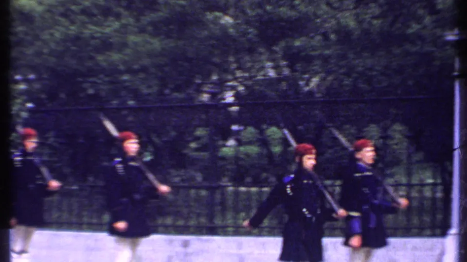 A line of uniformed men carrying rifles moves past a black iron fence, partly blurred by handheld 8mm camera movement.