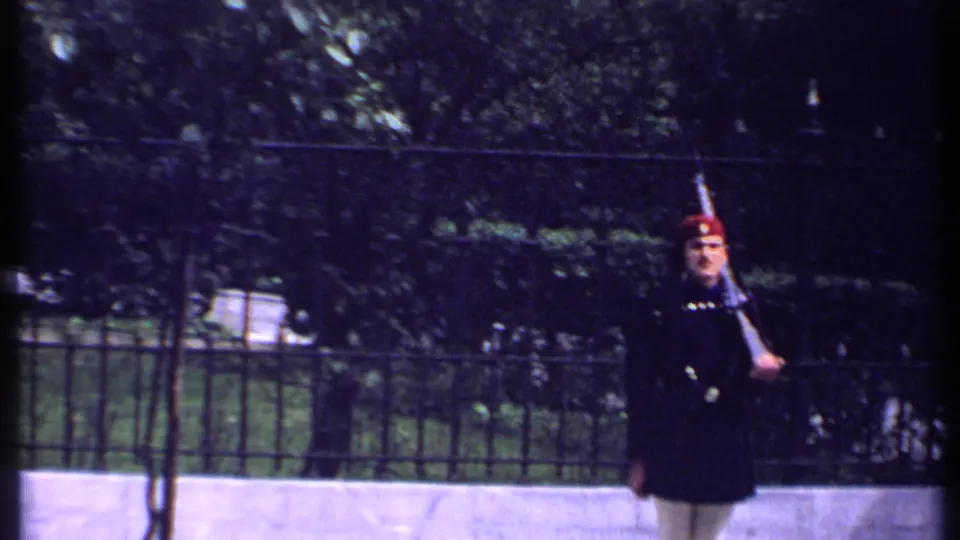 In 1969 Kenya footage, a uniformed guard in a red hat stands near a black iron fence with trees and hedges behind him.