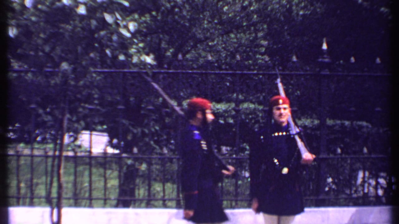 Two uniformed guards in red hats face each other near an iron fence.