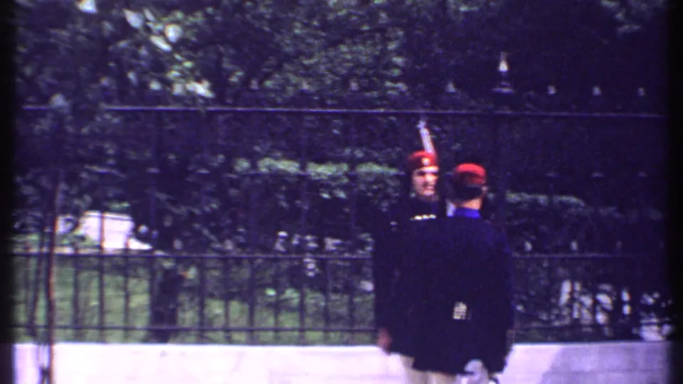 A rear-view guard faces a red-hatted guard near an iron fence, with garden greenery visible in the Kenya 1969 footage.