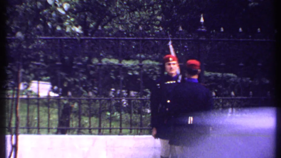 Two uniformed guards in red hats stand near a black iron fence, framed by trees and hedges in 1969 Kenya footage.