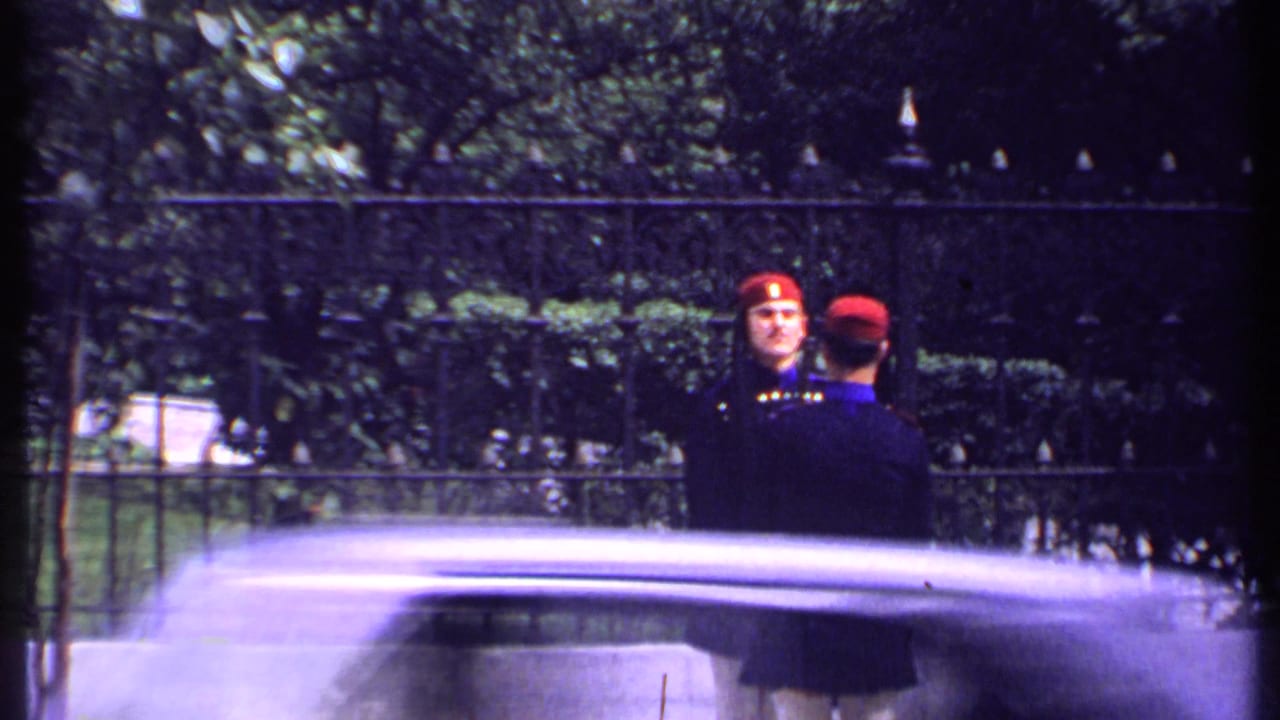 Two uniformed guards in red hats stand near an iron fence as a blurred vehicle passes in front.