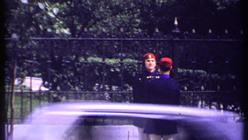 Kenya 1969 8mm frame of two ceremonial guards in dark uniforms and red hats near an iron fence, partly obscured by a passing vehicle.