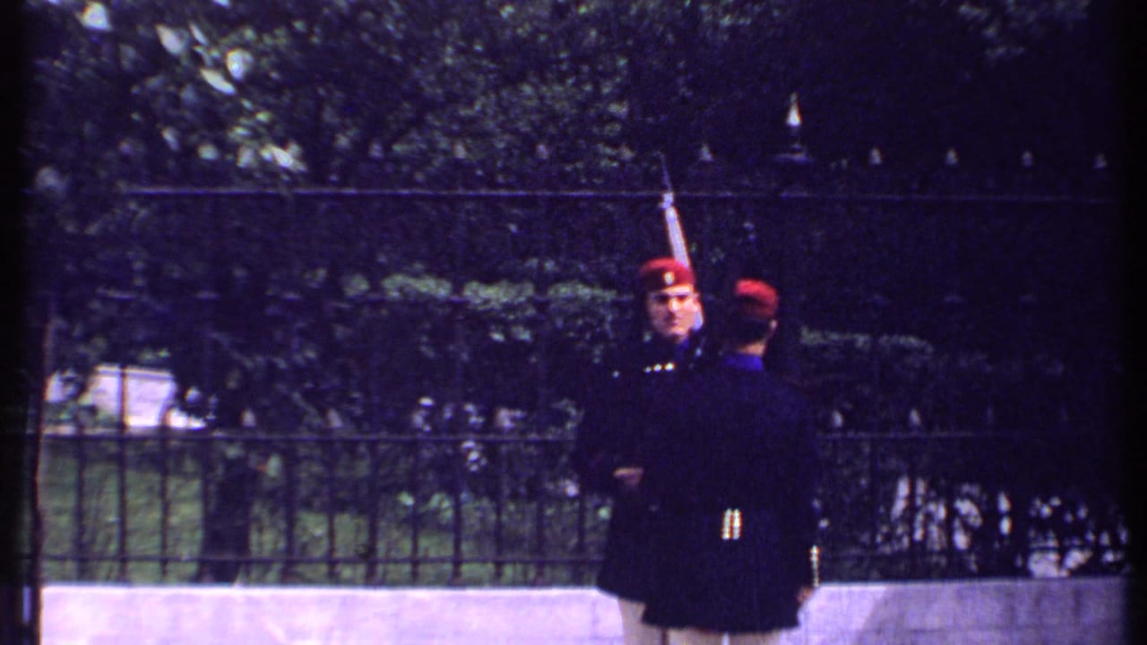 Two guards in dark uniforms and red hats face each other, one holding a rifle upright.