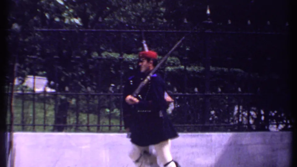 Kenya 1969 frame of a ceremonial guard in dark uniform and red hat moving with a rifle near an iron fence and trees.
