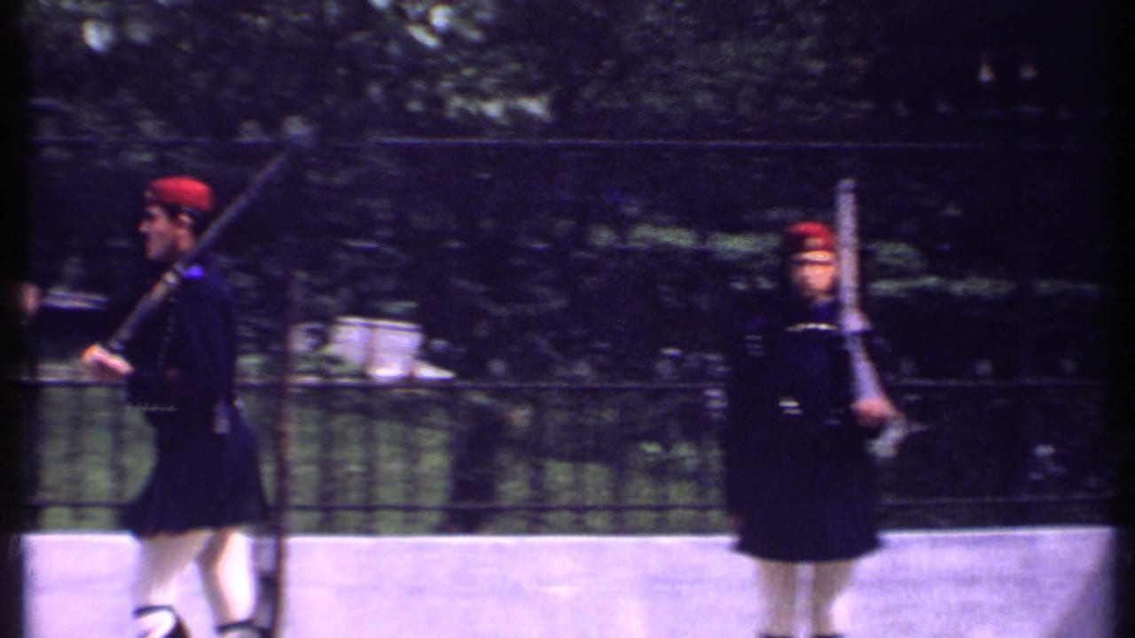 Uniformed soldiers in red hats march with rifles beside an iron fence.