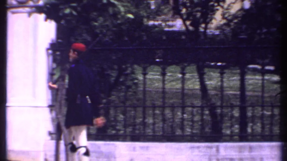 A soldier in dark ceremonial uniform and red hat is seen near pale masonry, with an iron fence and trees behind him in 1969 Kenya footage.