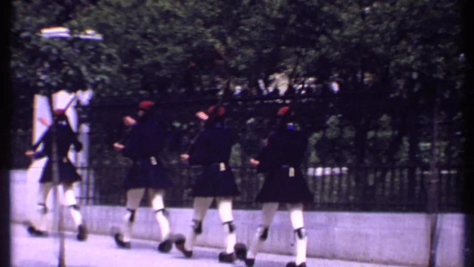 Several soldiers in dark uniforms and red hats march away with rifles along a paved path beside trees and iron fencing in Kenya, 1969.