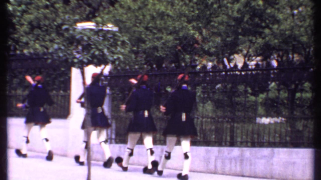 Four uniformed figures with rifles march beside a black iron fence.