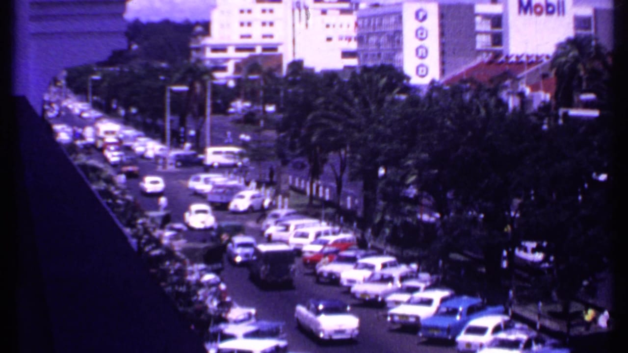 1969 Kenya home-movie frame showing dense car traffic, palm trees, and a prominent Mobil sign from an elevated street viewpoint.