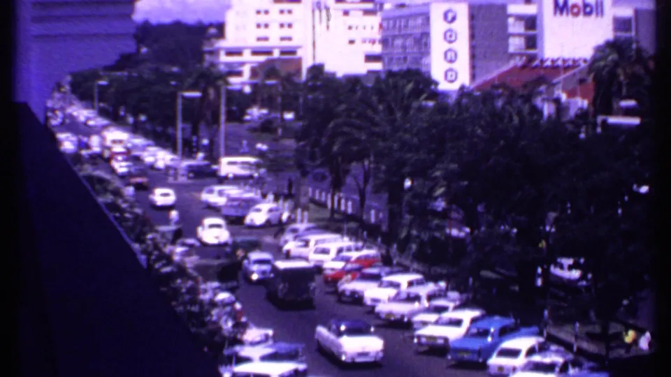 1969 Kenya home-movie frame showing dense car traffic, palm trees, and a prominent Mobil sign from an elevated street viewpoint.