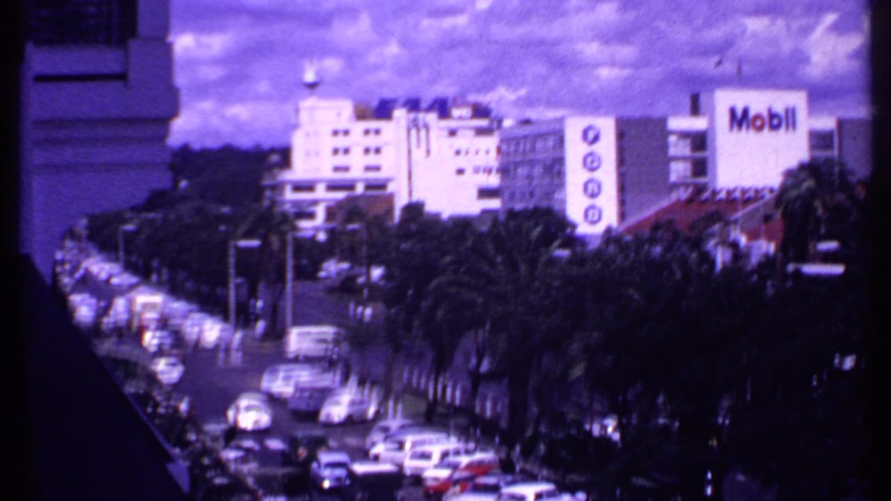 Palm-lined street with moving and parked cars, buildings, and Mobil signage beyond the road.