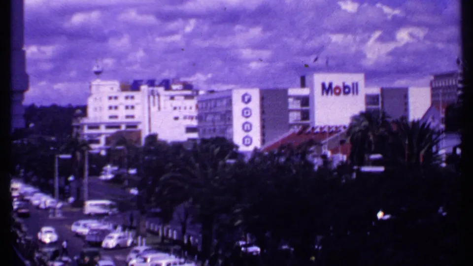 1969 Kenya home-movie view of a busy road with classic cars, palms, and Mobil and Ford signage on nearby buildings.