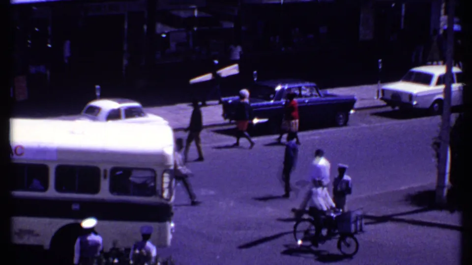 1969 Kenya urban street with the bus partly out of frame, pedestrians crossing, cars nearby, a cargo cyclist, and a traffic patrolman.