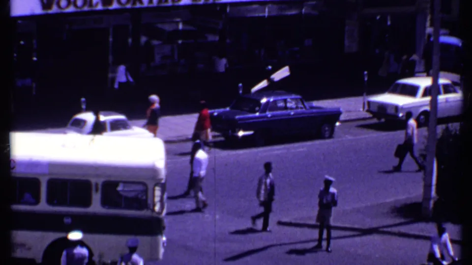 1969 Kenya street view showing classic sedans, pedestrians, a partial bus, storefront signage, and a uniformed patrolman observing traffic.