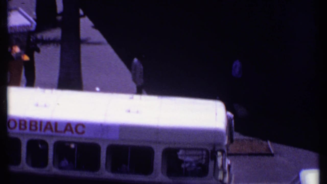 Man in a white suit crosses between a station wagon and a bus with ROBBIALAC lettering.