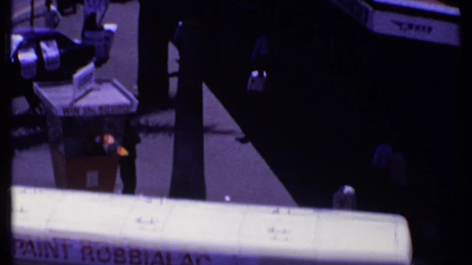 1969 Kenya frame showing a street corner, newsstand, bus roof, pedestrians, and deep shadows from nearby trees or palms.