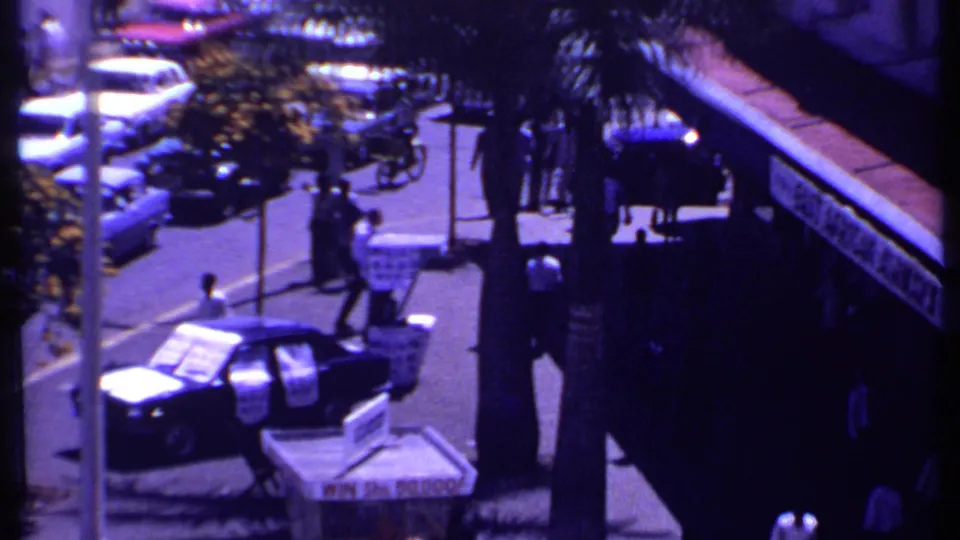 1969 Kenya home-movie view from above a busy street with cars, pedestrians, palm trees, signage, and a dark storefront canopy.